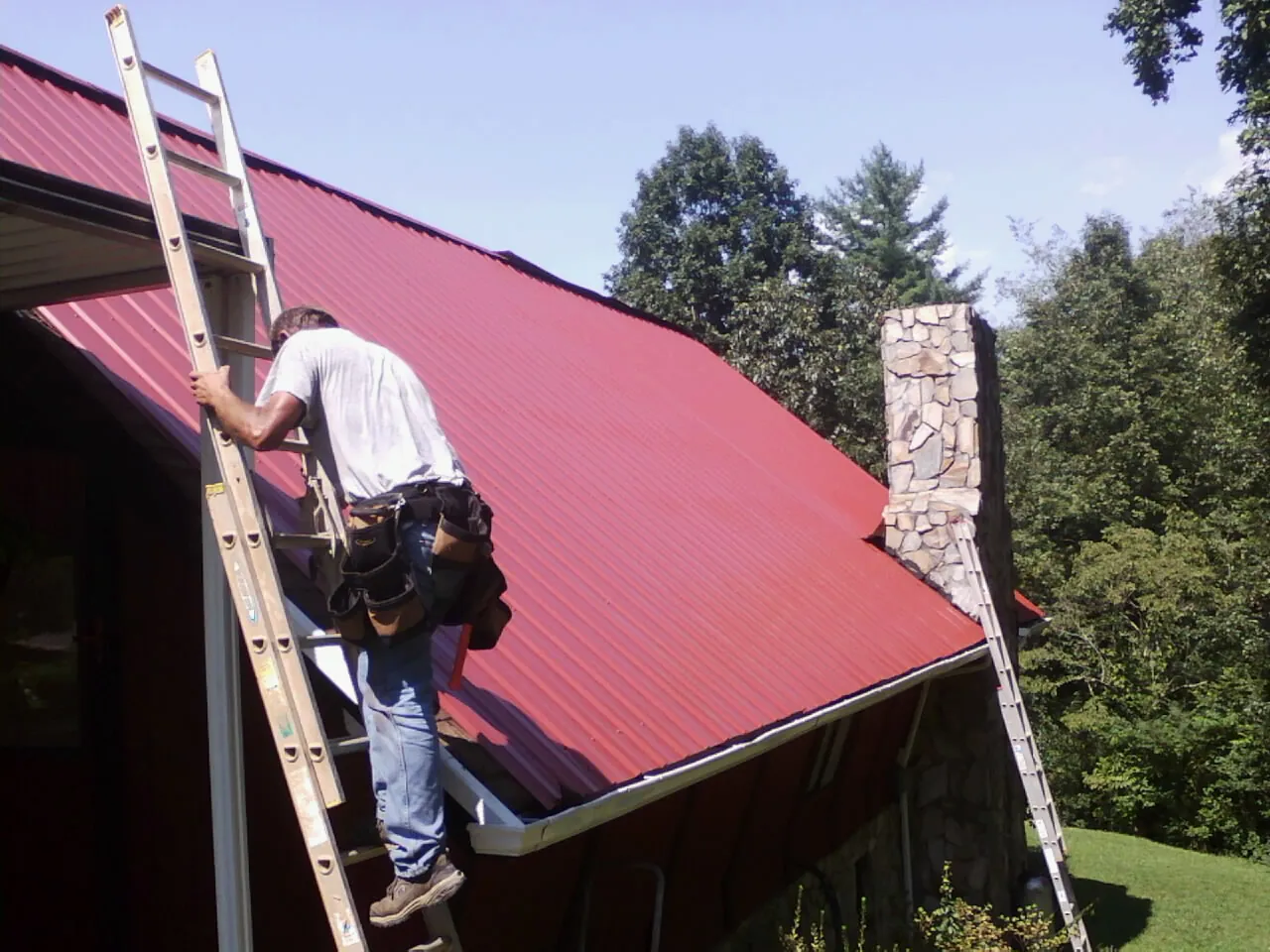 Roofer on a ladder beside a red metal roof and stone chimney — Soffit & Fascia Repair by Roofing Brothers in Beechgrove